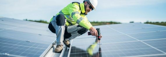 Male worker on a roof using drill to fix solar panel.